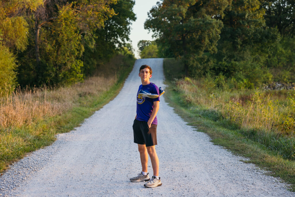 Kansas City senior boy holding cross country shoes on rural dirt road during senior session